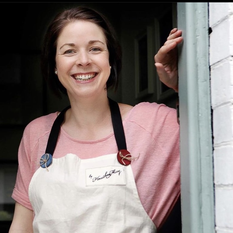 Heather Brown leaning on a doorway wearing her By Heather May apron
